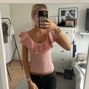Rosa volangtopp med smock - Supersöt rosa topp med smockat tyg och bred volang runt ringningen. Toppen är ärmlös och har en croppad passform som sitter tajt mot kroppen. Perfekt för dig som gillar romantiska detaljer och en feminin look.