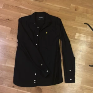 Svart skjorta från Lyle & Scott S - Svart skjorta från Lyle & Scott med klassisk krage, vita knappar och bröstficka med gul broderad logga. Skjortan är långärmad och har en stilren, enkel design som passar till många olika looks.