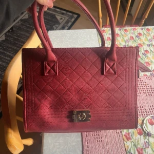 Röd quiltad handväska från Erich - Snygg röd handväska med quiltat mönster och guldfärgad låsdetalj framtill. Väskan har två rejäla handtag, en avtagbar axelrem och dragkedja baktill. Materialet är skinnimitation och insidan är rymlig med extra fack. Perfekt för dig som gillar stilrena accessoarer. Oanvänd