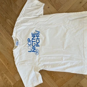 Vit t-shirt från LOOP Nicotine PCHS - Säljer en vit t-shirt från LOOP Nicotine PCHS i storlek L. T-shirten har blå tryck framtill med cool grafisk text och klassisk rund hals. Modellen är kortärmad och gjord i mjuk bomull, perfekt för en chill och avslappnad stil. Kan bara fås av säljare, sällsynt