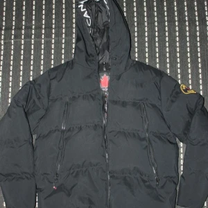 Svart pufferjacka Zavetti Canada XL - Svart pufferjacka från Zavetti Canada i storlek XL. Jackan har huva med tryckt logga, två vertikala dragkedjefickor fram och emblem på ärmen. Materialet är syntet och polyester, perfekt för kalla dagar. Snyggt broderad lönnlogga på insidan.