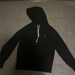 Svart hoodie från Polo Ralph Lauren - Svart hoodie från Polo Ralph Lauren med vit snörning och rött broderat logomärke på bröstet. Tröjan har huva och långa ärmar, tillverkad i mjuk bomull. Perfekt för chill dagar och enkel att matcha med jeans eller joggers.