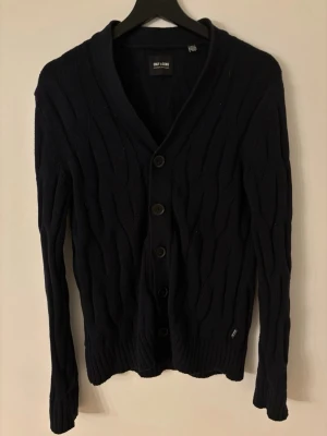 Mörkblå kofta från Only & Sons - Snygg mörkblå kofta från Only & Sons med kabelstickat mönster och fem knappar framtill. Koftan har långärmad passform och ribbade muddar vid ärmslut och nederkant. Perfekt att slänga på över en t-shirt eller hoodie för en chill look.