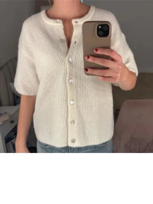 Vit stickad kofta med kort ärm - Superfin vit stickad kofta med korta ärmar och rund hals. Koftan har ribbad struktur och knäpps med pärlemorknappar framtill. Passformen är normal och materialet känns mjukt och fluffigt, perfekt att slänga över en topp eller linne. Har några få noppror som inte syns och är lätta att få bort