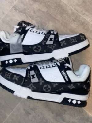 Louis Vuitton sneakers svart/vit - Säljer ett par Louis Vuitton sneakers i svart och vitt med klassiskt monogrammönster. Skorna har snörning, platt sula och rund tå. Materialet är en mix av skinn och textil, med tydliga LV-detaljer på sidan och tungan. Perfekta för dig som gillar lyxiga streetwear-vibes.