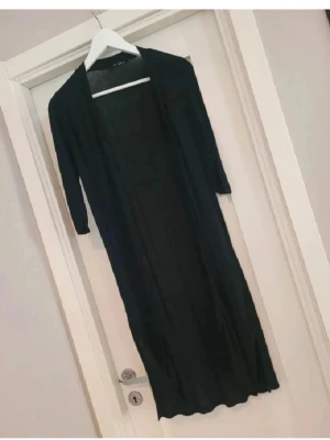 Svart lång kofta med trekvartsärm - Lång svart kofta med trekvartsärm, perfekt att slänga över en topp eller klänning. Enkel och stilren design utan knappar eller dragkedja. Tunnare material som ger ett snyggt fall och passar till många olika outfits.