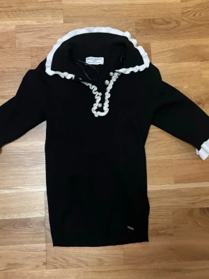 Svart stickad kofta från Holly & Whyte - Svart stickad kofta med vit volangkant vid kragen och knäppning med pärlknappar. Långärmad modell med ribbad struktur och vit detalj vid ärmslut. Perfekt för dig som gillar klassiska detaljer med en twist.