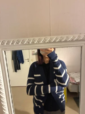 Marinblå randig kofta från Manguun - Säljer en marinblå stickad kofta från Manguun med vita horisontella ränder. Koftan har v-ringad hals, långa ärmar och knäpps med en knapp framtill. Perfekt för dig som gillar chill och stilren look.