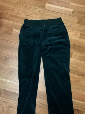 Mörkgröna mjukisbyxor Juicy Couture S - Säljer ett par mörkgröna mjukisbyxor från Juicy Couture i storlek S. Byxorna har bred resår med snörning i midjan, två stora fickor fram och ett broderat Juicy Couture-logo på ena fickan. Materialet är mjuk sammet som känns lyxigt och skönt.