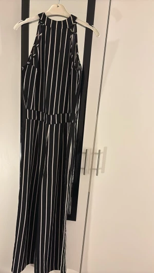 Svartvit randig byxdress - Snygg svartvit byxdress med vertikala ränder och hög hals. Byxdressen har vida ben och knäppning i nacken. Materialet känns lätt och luftigt, perfekt för dig som gillar en cool och trendig look. Passar dig som vill sticka ut med stilrena detaljer.