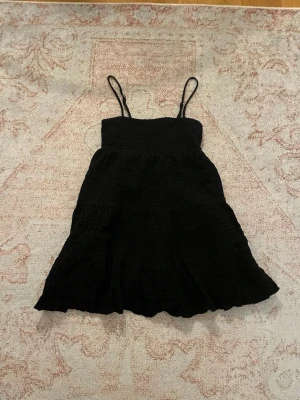 Svart kort klänning från Zara - Supersöt svart klänning från Zara med smockad överdel och tunna axelband. Klänningen har en skrynklig textur. Perfekt för dig som gillar enkel men snygg stil. Passar dig som vill ha något bekvämt och trendigt.