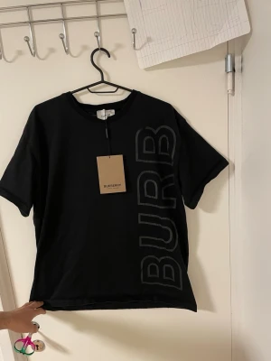 Svart Burberry t-shirt med tryck - Säljer en svart t-shirt från Burberry med stort, ton-i-ton tryck som sträcker sig vertikalt över både fram- och baksidan. T-shirten har rund hals och korta ärmar, tillverkad i mjuk bomull för skön känsla. Perfekt för dig som gillar stilrena designer med tydlig logga.Liknande modell som originala.