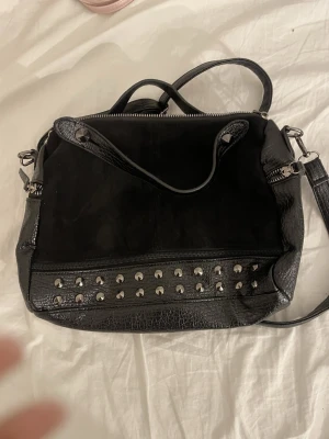 Svart axelväska med nitar - Cool svart axelväska i en mix av mocka och skinnimitation. Väskan har både handtag och justerbar axelrem, samt snygga silverfärgade nitar på framsidan och flera dragkedjor. 