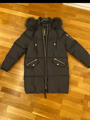 Svart pufferjacka från River Island - Säljer en svart pufferjacka från River Island med fluffig fuskpäls på huvan, långa ärmar och flera fickor med dragkedja och knappar. Jackan har en snygg oversized look och är perfekt för kalla dagar. Materialet är polyester och jackan stängs med dragkedja framtill.