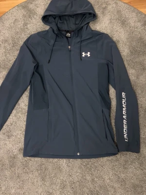 Under Armour Vindjacka - blå/vit - Snygg helt oanvänd Under armour jacka i S. Den har en väldigt unik design som är svår att få tag på där det står ”under armour” på armen. Riktigt fet. Kan lösa gratis frakt och sänka priset vid snabb affär.