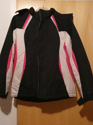 Vinterjacka - Säljer en sportig vinterjacka från George i svart, rosa och vitt. Jackan har huva med mjukt fleecefoder, dragkedja framtill och justerbara muddar. Snygga färgblock längs sidorna och insidan av huvan är rosa. Perfekt för kalla dagar! Har fläckar.