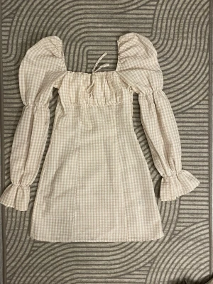 Beige rutig klänning från H&M - Säljer en beige och vit rutig kortklänning från H&M med puffiga långärmar och volang vid ärmslut. Klänningen har en söt knytning vid halsen och är tillverkad i ett lätt bomullsmaterial. Använd få gånger! 