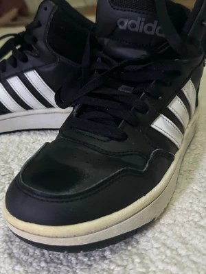 Svarta Adidas sneakers med vita ränder - Säljer ett par svarta Adidas sneakers med klassiska vita ränder på sidorna och vit sula. Skorna har snörning och hög modell som ger extra stöd runt ankeln. Adidas-logga syns både på plösen och hälen. Materialet är en mix av skinn och syntet.