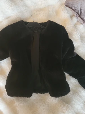 Svart pälsjacka  - Säljer en svart pälsjacka från H&M med rund hals och långärmade ärmar. Jackan har en mjuk och fluffig yta som ger en lyxig känsla och är perfekt för att hålla sig varm under kyliga dagar. Enkel och stilren design utan synliga knappar eller dragkedja.