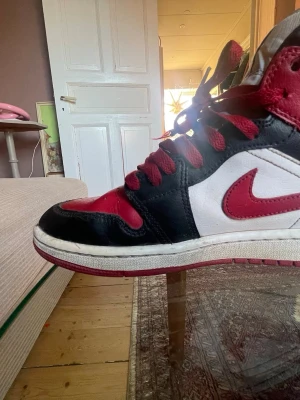 Nike Air Jordan 1 High röd/svart/vit - Säljer ett par Nike Air Jordan 1 High med klassisk färgblockning i rött, svart och vitt. Skorna har röd swoosh, röda snören och perforerad tå. Ovandel i skinn och hög siluett som ger en ikonisk look. Perfekt för dig som gillar streetwear och sneakers.