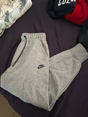 Gråa mjukisbyxor från Nike ( Niketech ) - Säljer ett par helt nya grå mjukisbyxor från Nike i storlek  XS ( 34 ) men passar också bra för de som har S. De köptes för 1200kr men säljs för 700kr för att jag vill bli av med de. De har svart logga på vänster ben. De har har snörning i midjan, ribbade muddar vid bensluten och flera fickor, inklusive en med dragkedja på sidan. Perfekta för chill eller träning, jätte sköna och stilrena. Byxorna har ALDRIG använts. Har inte möjlighet att visa hur de sitter på eftersom att de är för små för mig.