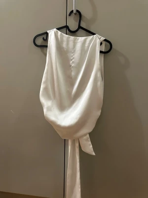 Vit topp med rosett i bak - Säljer en elegant vit topp i glansigt satinliknande material. Toppen är ärmlös med breda axelband och har en djup v-ringning i bak. Den knyts med en stor rosett i midjan för en snygg siluett.