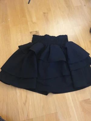 Svart volangkjol från H&M - Säljer en svart kortkjol från H&M med bred smock i midjan och flera lager volanger som ger en snygg och trendig look. Kjolen är gjord i ett mjukt syntetmaterial och passar perfekt för dig som gillar att sticka ut med coola detaljer.