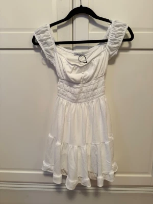 Vit kortklänning från Bershka - Supersöt vit kortklänning från Bershka i storlek XS. Klänningen har offshoulder-ärmar, smockad midja och volangdetaljer längst ner. Materialet är mjukt och lätt, perfekt för varma dagar. Passformen är normal och klänningen har en romantisk vibe.