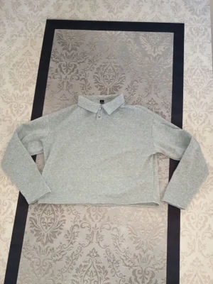 Grå pikétröja från UrbanChill - Snygg grå pikétröja från UrbanChill med klassisk krage och två knappar framtill. Tröjan har lång ärm och är croppad i modellen, vilket ger en modern och avslappnad vibe. Perfekt för dig som gillar enkel och stilren design.