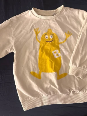 Vit tröja med Bii-tryck från Kappahl - Säljer en gul långärmad tröja från Kappahl med ett stort gult Bii-tryck framtill. Tröjan har ribbade muddar vid hals, ärmslut och nederkant. Materialet är mjuk bomull, perfekt för chill dagar. Rolig design som sticker ut!