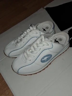 Vita sneakers från Kangaroos strl 35 - Snygga vita sneakers från Kangaroos med ljusblå detaljer och klassisk logga på sidan. Skorna har snörning, rund tå och en sula i gummi med ljusblå inslag undertill. Ovandelen är i syntetmaterial och insidan är fodrad med textil för extra komfort. Helt nya men har några skador pga att de varit intryckta se bilder