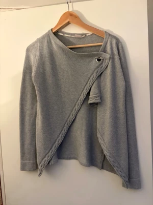 Grå omlottkofta från Object XS - Säljer en grå omlottkofta från Object i storlek XS. Koftan har lång ärm, stickad struktur och snygg snörning med metallring vid axeln. Dekorativ flätad kant längs omlotten ger extra stilpoäng. Perfekt för lager på lager under kyliga dagar.