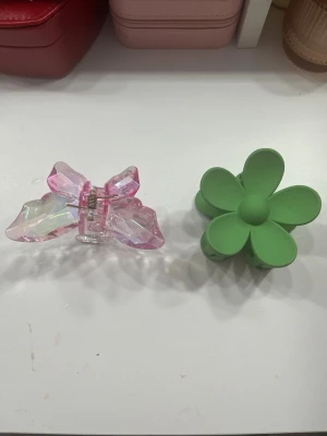 Grön blomformad och rosa fjärils hårklämma - Två hårklämmor säljes! Ena är grön och formad som en blomma med rundade blad, den andra är transparent rosa och glittrig, formad som en fjäril med facetterade vingar. Båda är i plast och har starka fjädrar för att hålla håret på plats.