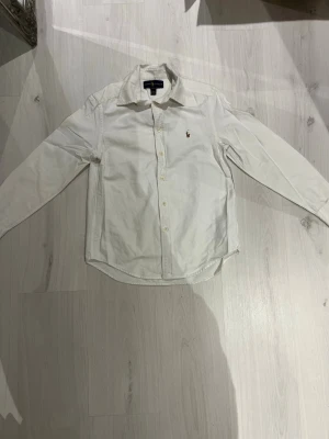 Vit skjorta från Ralph Lauren - Klassisk vit skjorta från Ralph Lauren med broderad logga på bröstet. Skjortan har lång ärm, knappar framtill och en snygg krage. Tillverkad i mjuk bomull som känns skön mot huden. Perfekt för dig som gillar stilrena och tidlösa plagg.