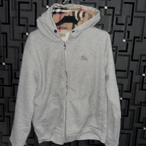 Grå Burberry hoodie med dragkedja M - Säljer en grå hoodie från Burberry i storlek M. Tröjan har klassisk dragkedja, huva med det ikoniska rutiga Burberry-mönstret på insidan och två fickor framtill. Små fläckar som kanske går bort i tvätten 