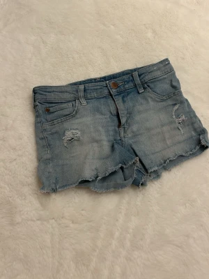 Ljusblå denimshorts från H&M - Säljer ett par ljusblå denimshorts från H&M med slitna detaljer och fransig kant. Klassisk femficksmodell med knapp och dragkedja. Perfekta för varma dagar och ger en avslappnad vibe. Storlek 140 men passar även uppemot 155.🥰