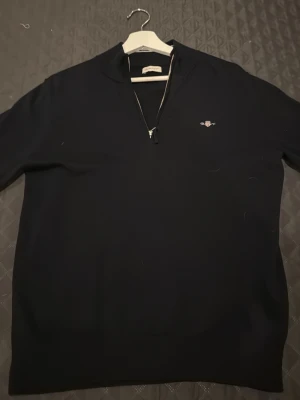 Svart half zip tröja från GANT - Svart långärmad tröja från GANT med half zip framtill och broderad logga på bröstet. Tröjan har en stilren design och är gjord i mjukt material som passar perfekt till kyligare dagar. Klassisk passform och diskret krage.