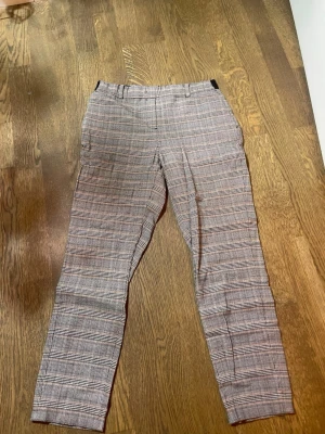 Rutig kostymbyxa med smal passform - Snygga kostymbyxor med rutig design i svart, vit och orange. Byxorna har smal passform, bälteshällor och klassiska fickor både fram och bak. Materialet är mjukt. Perfekt för dig som gillar stilrena och trendiga mönster.