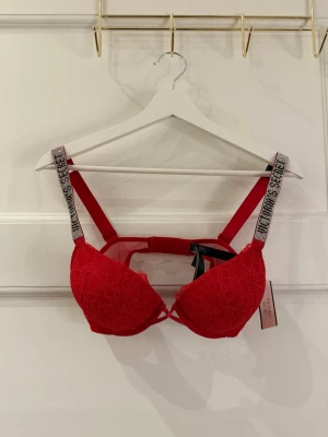 Röd spets-BH från Victoria's Secret - Säljer en röd push-up BH från Victoria's Secret med spetsdetaljer och glittriga axelband. Kuporna har ett snyggt spetsmönster och axelbanden är dekorerade med strass. BH:n har justerbara band och hakknäppning bak. Den är helt oanvänd med lapparna kvar.