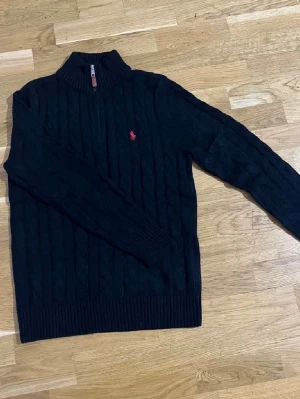Svart stickad tröja Polo Ralph Lauren - Svart stickad tröja från Polo Ralph Lauren med halv dragkedja och ribbad krage. Klassisk kabelstickad design och röd broderad logga på bröstet. Långärmad och med ribbade muddar för extra komfort. Perfekt för dig som gillar stilrena och tidlösa plagg.