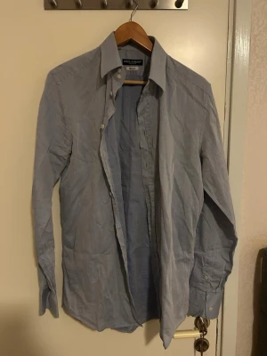 Ljusblå skjorta Dolce & Gabbana Gold - Snygg ljusblå skjorta från Dolce & Gabbana Gold med klassisk krage och knappar framtill. Skjortan har lång ärm och är tillverkad i ett tunt, vävt bomullstyg som känns fräscht och stilrent. Perfekt för dig som gillar italiensk design och vill ha en clean look. Storleken är 40 passar jättebra till dom som har storlek M 