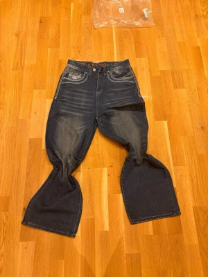 Rock Revival bootcut jeans blå - Säljer ett par blå bootcut jeans från Rock Revival med kontrasterande vita sömmar och broderade detaljer på fickorna. Jeansen har låg midja och klassisk femficksdesign med coola loggor och slitningar för extra edge.