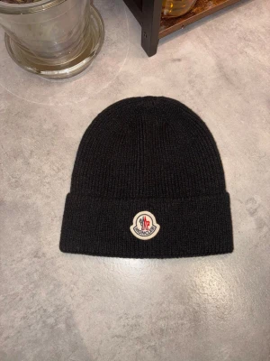 Svart Moncler mössa i ull - Svart ribbstickad mössa från Moncler med klassisk uppvikt kant och broderad logotyp framtill. Tillverkad i mjuk ull som håller dig varm och snygg under kyliga dagar. Perfekt accessoar för dig som gillar stilrena detaljer och exklusiva märken.