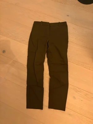 Bruna kostymbyxor från H&M, strl 36 - Snygga bruna kostymbyxor från H&M i storlek 36. Byxorna har klassisk rak passform med hällor för bälte och stängs med hake och dragkedja. Materialet är mjukt och känns bekvämt mot huden, perfekt för en clean och stilren look.