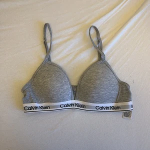 Grå Calvin Klein topp med logoband - Säljer en grå topp från Calvin Klein med justerbara axelband och ett vitt elastiskt band med svart logotyp längs nederkanten. Mjuk och skön bomullskänsla, perfekt för chill dagar. Enkel design med sportig vibe.🩷
