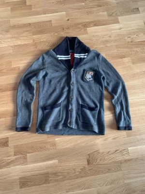 Grå kofta från Morris med emblem - Snygg grå kofta från Morris University Collection med mörkblå detaljer på krage och fickor. Koftan har ett broderat emblem på bröstet, knappar framtill och ribbade muddar. Perfekt för dig som gillar klassisk stil med en preppy touch.