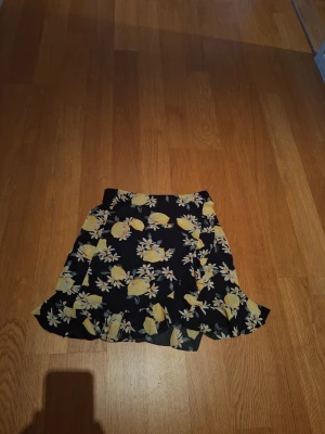 Blommig kortkjol från Gina Tricot - Supersöt kortkjol från Gina Tricot med svart botten och gult blommönster. Kjolen har volangkant nertill och omlott-look som ger en extra snygg touch. Perfekt för dig som vill ha en lekfull och trendig stil.