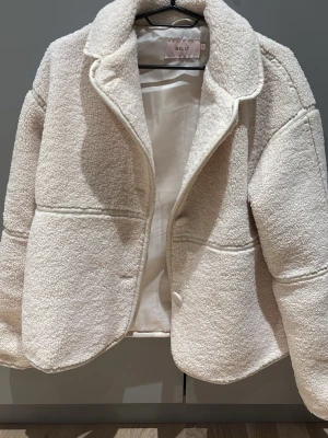 Beige teddyjacka från Nelly - Mysig teddyjacka i beige från Nelly. Jackan har tre knappar framtill och markerade sömmar som ger en cool look. Perfekt för att hålla sig varm och samtidigt se stylish ut.
