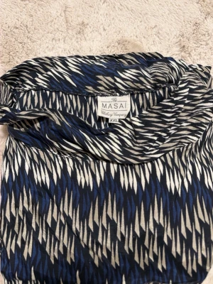 Mönstrad klänning från Masai XXL - Säljer en snygg klänning från Masai i storlek XXL med ett coolt grafiskt mönster i blått, svart och beige. Klänningen har lång ärm och en bred krage som ger en avslappnad look. Materialet känns lätt och skönt mot huden.