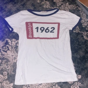 Vit t-shirt Brooklyn 1962 Pull&Bear - Vit t-shirt från Pull&Bear med trycket 'Brooklyn 1962' i rött och svart på bröstet. Rund hals med marinblå kant och matchande blå detaljer vid ärmslut. Klassisk kortärmad modell i mjuk bomull, perfekt för en chill och avslappnad stil.